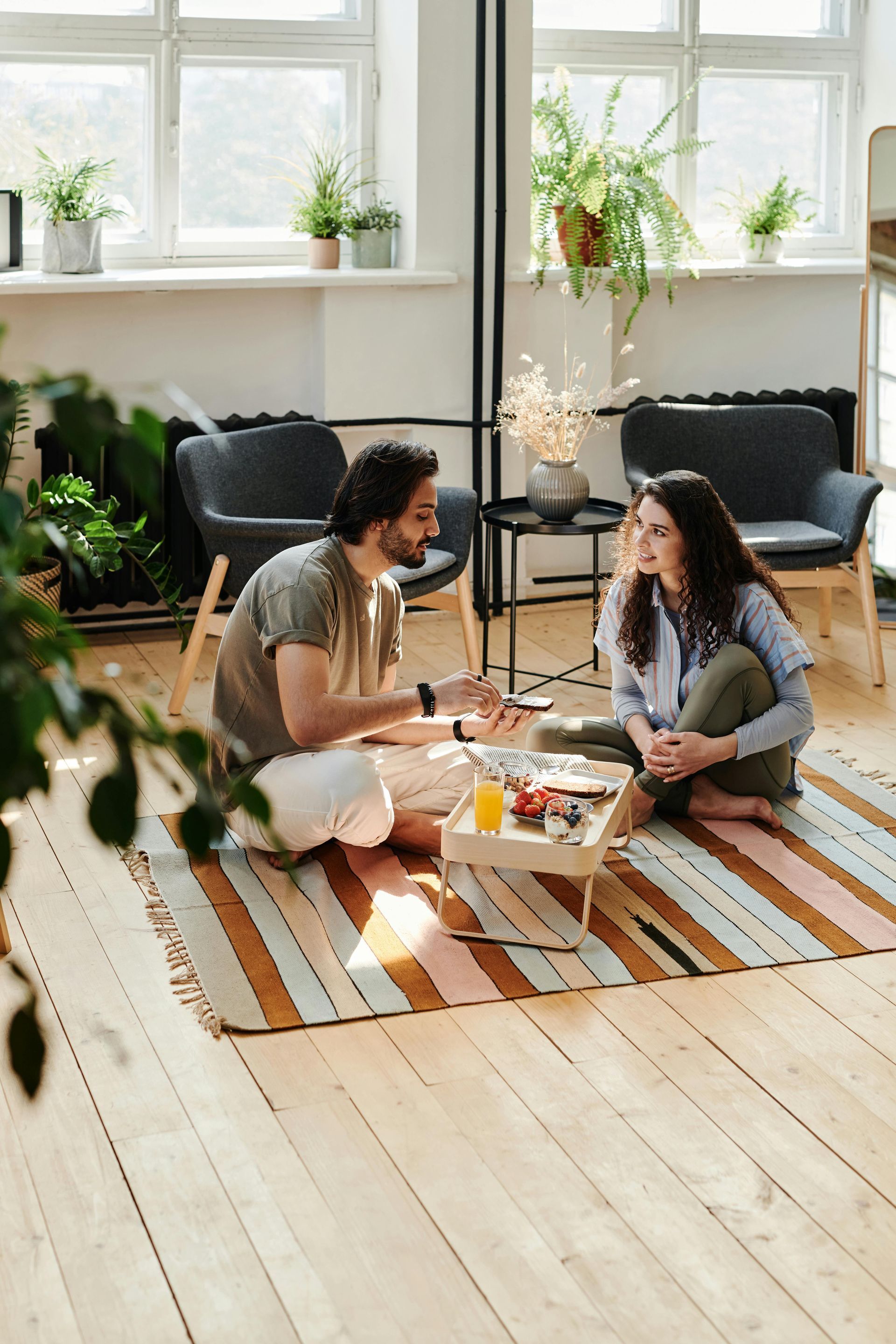 Man and woman sit on rug in a sunny room, talking and eating from a tray.