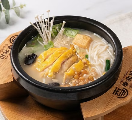 Black bowl of noodle soup with sliced chicken, mushrooms, and tofu on a wooden trivet