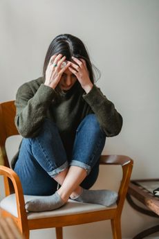 Woman sitting on a chair, hugging knees, hands on head, looking down, appearing stressed.