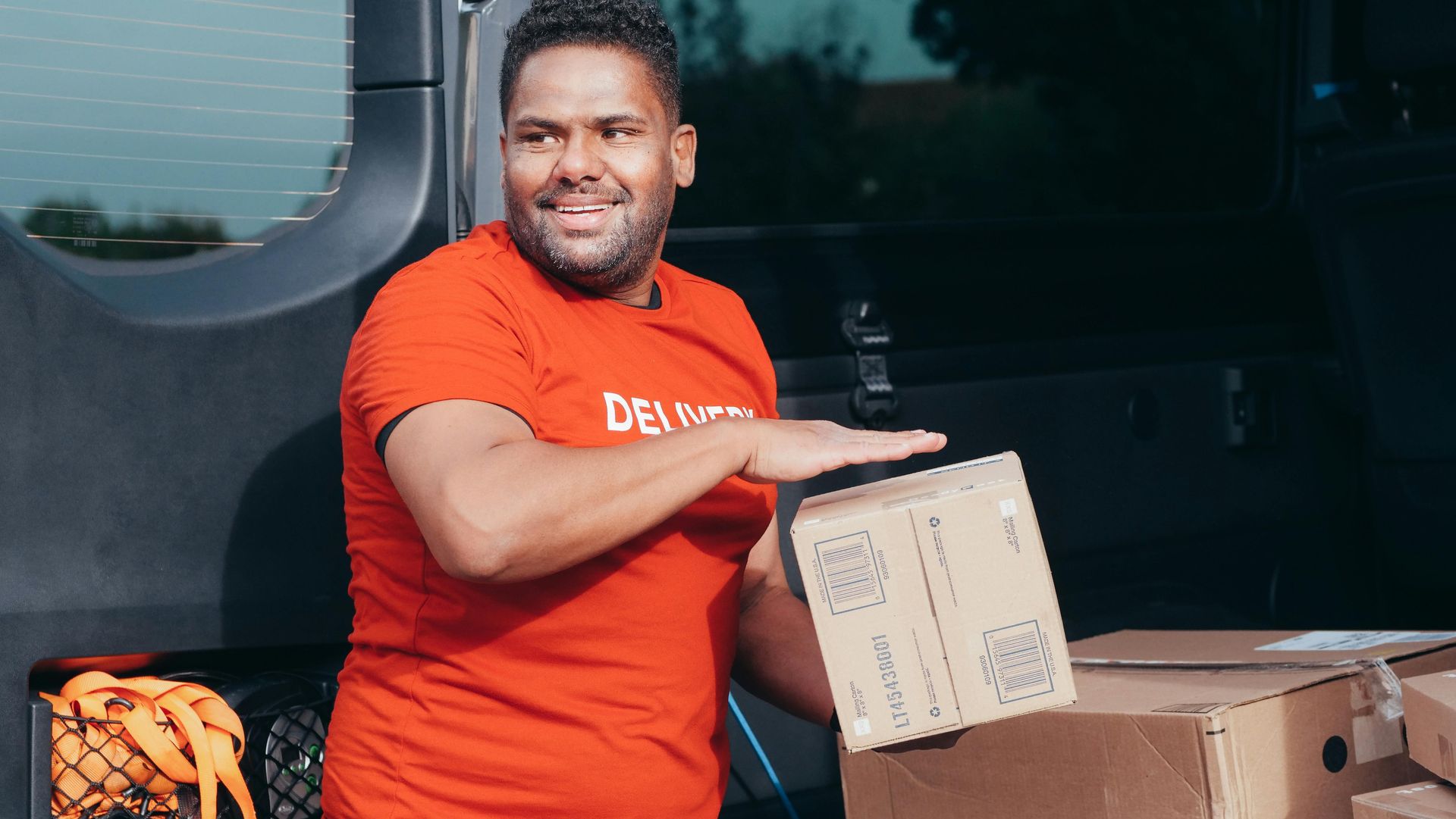 Delivery person in red shirt smiles while reaching for box in delivery van.