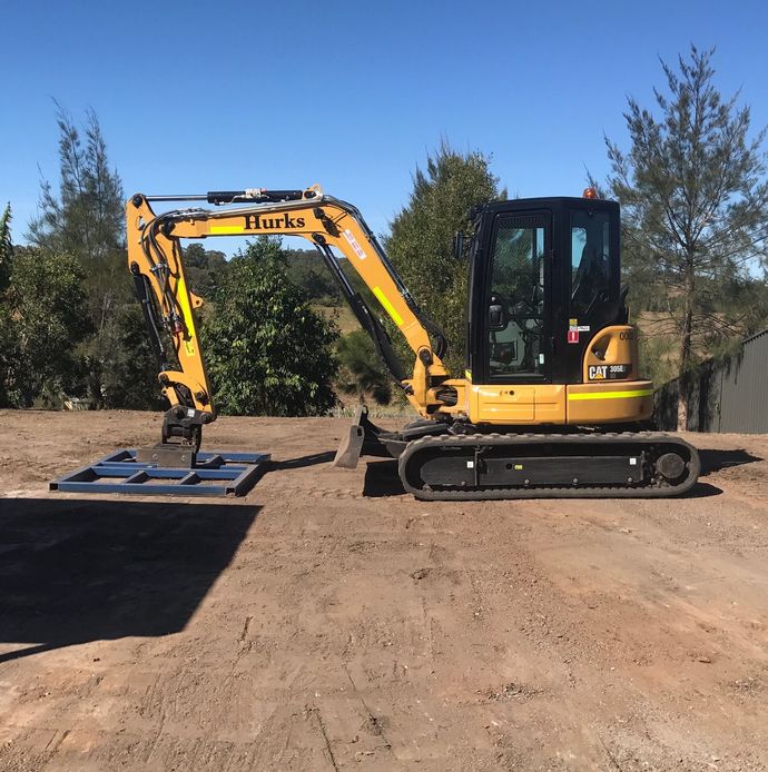 Yellow excavator on a dirt lot, positioned beside a tree and black platform under a clear blue sky.  — Jensen Welding & Fabrication in Chevallum, QLD