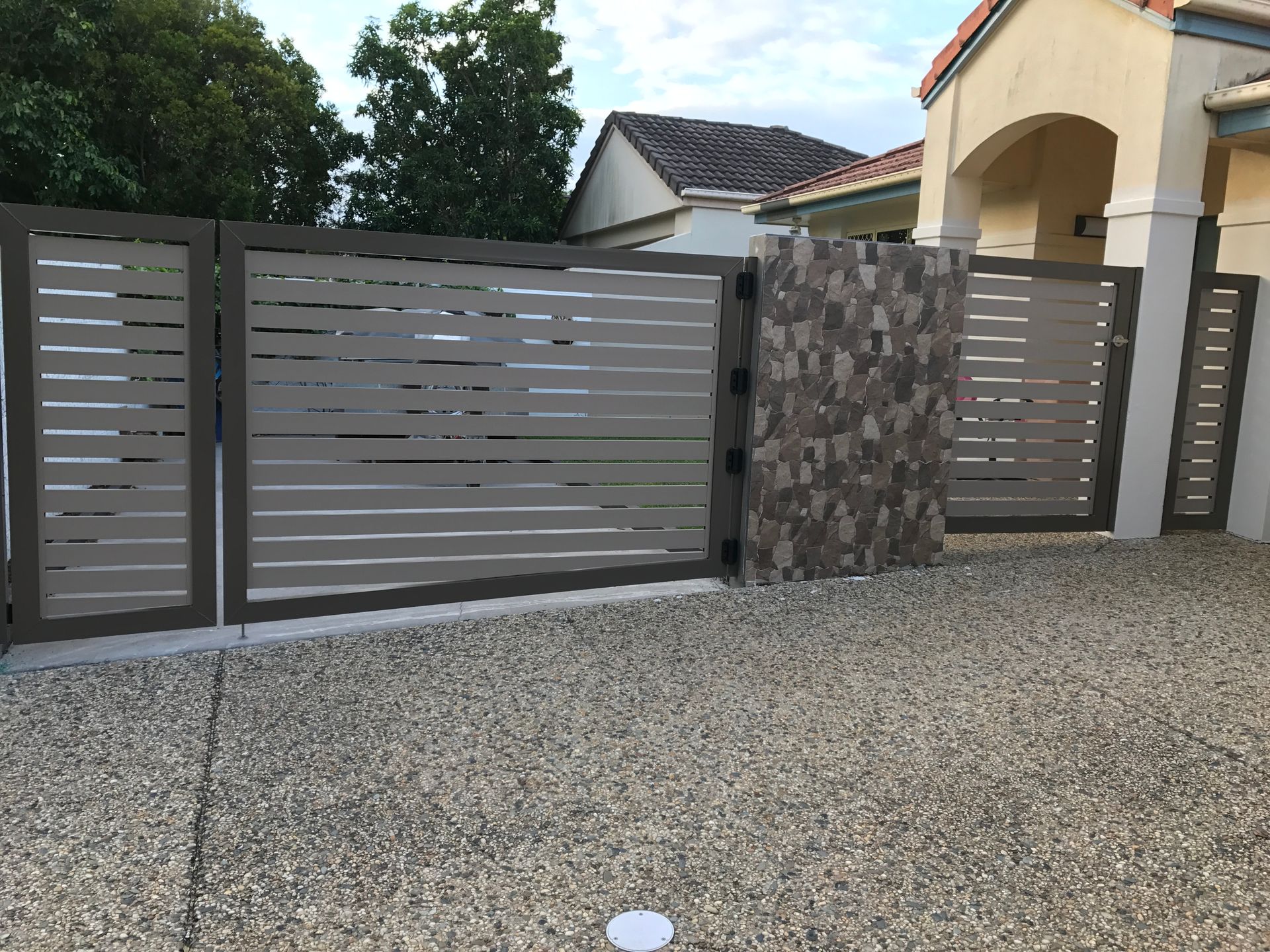 Modern gray metal gate and fence in front of a house driveway, with stone column and gravel ground  — Jensen Welding & Fabrication in Chevallum, QLD