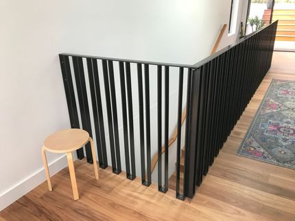 Black stair railing with vertical slats beside a small wooden stool in a bright hallway — Jensen Welding & Fabrication in Chevallum, QLD