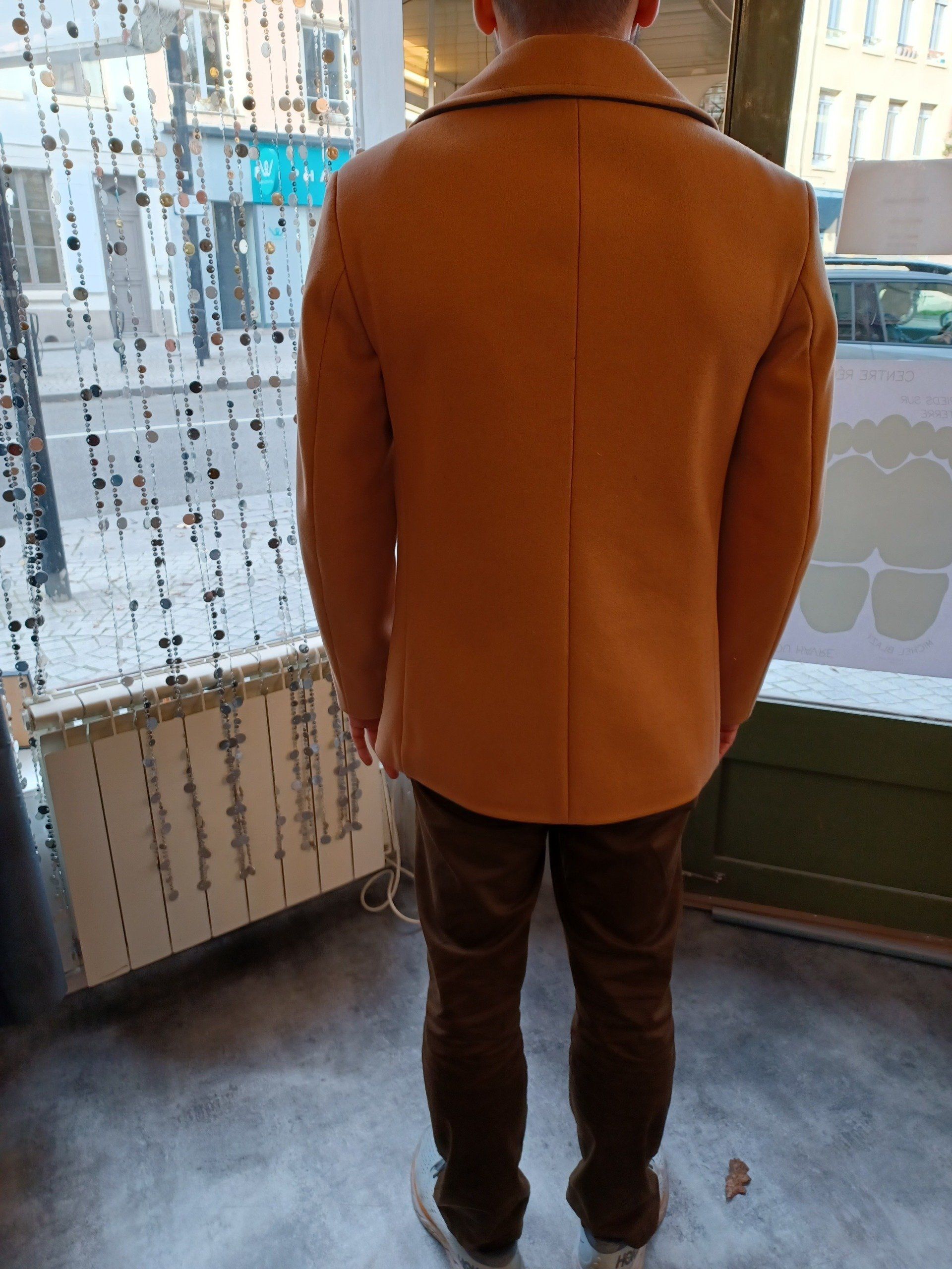 Un homme portant une veste marron se tient devant une fenêtre.