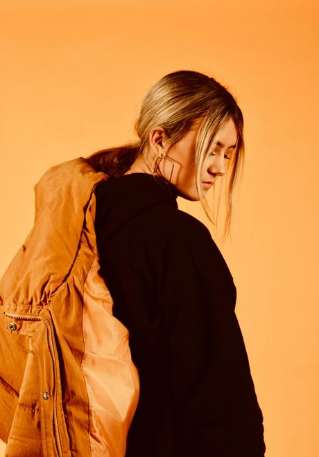 Une femme portant un pull noir et une veste marron se tient devant un fond orange.