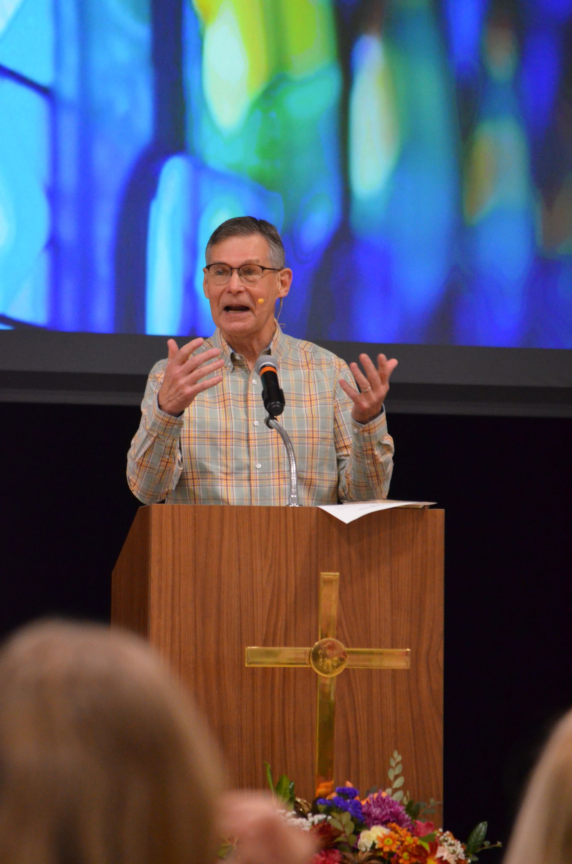 Man speaking at a podium with a cross on it; colorful background.