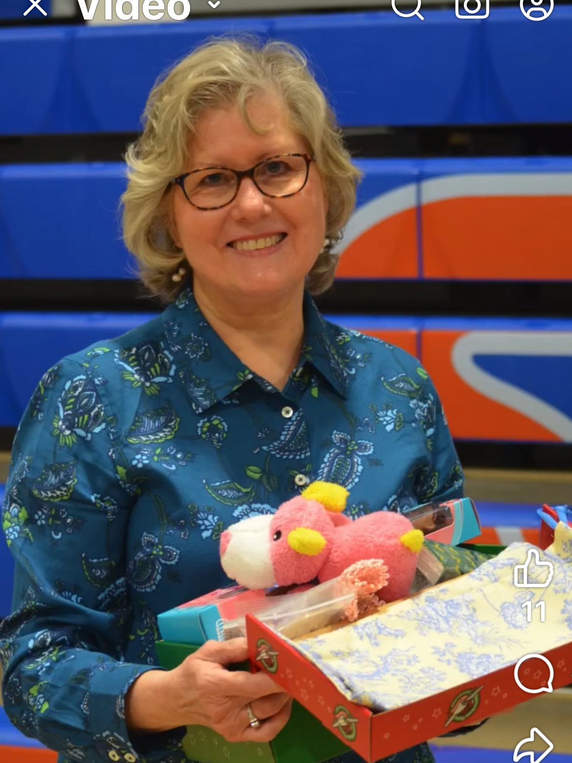 A woman wearing glasses is holding a stuffed animal and a box of toys.