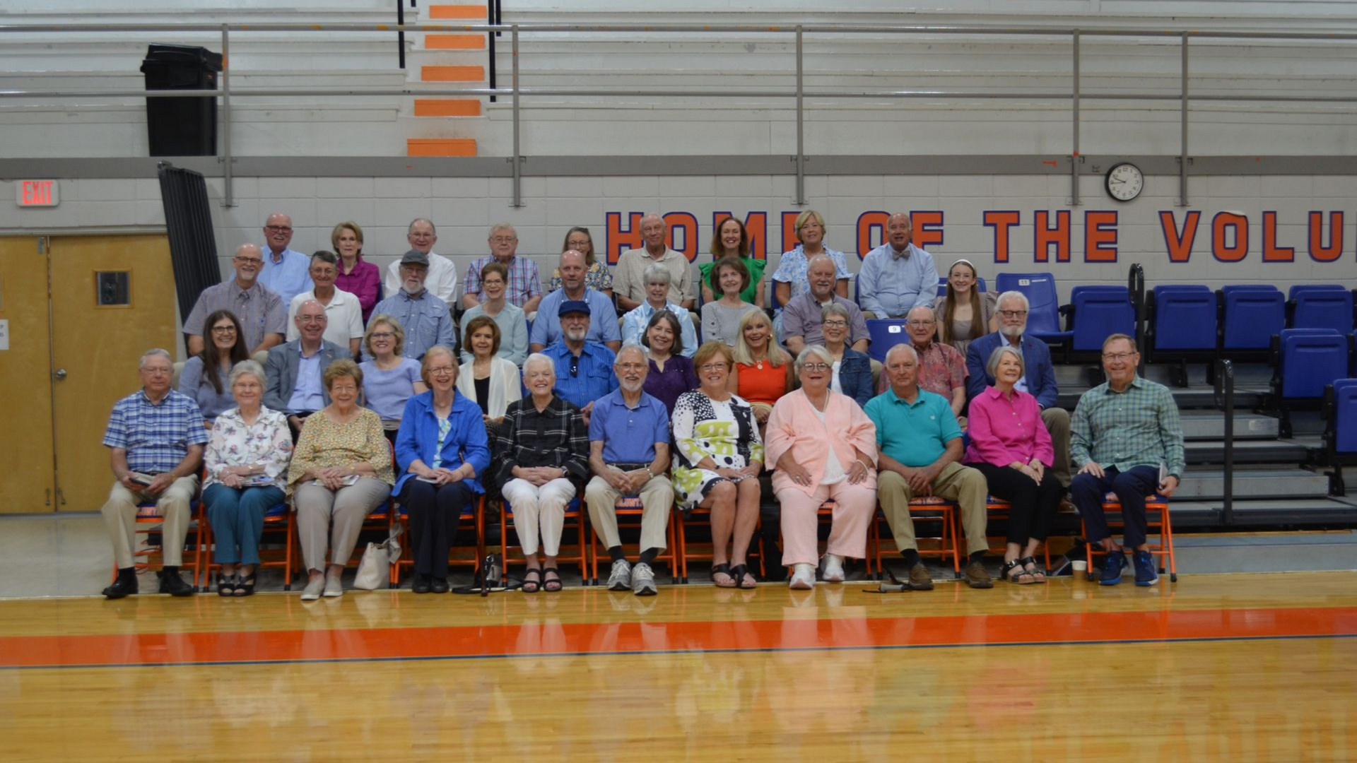 Group of people sitting and standing in a gymnasium, orange and blue accents, banner: 