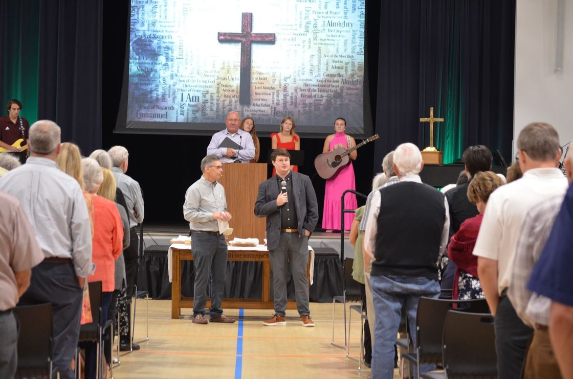Church service: People facing stage with cross. A man speaks into a microphone.