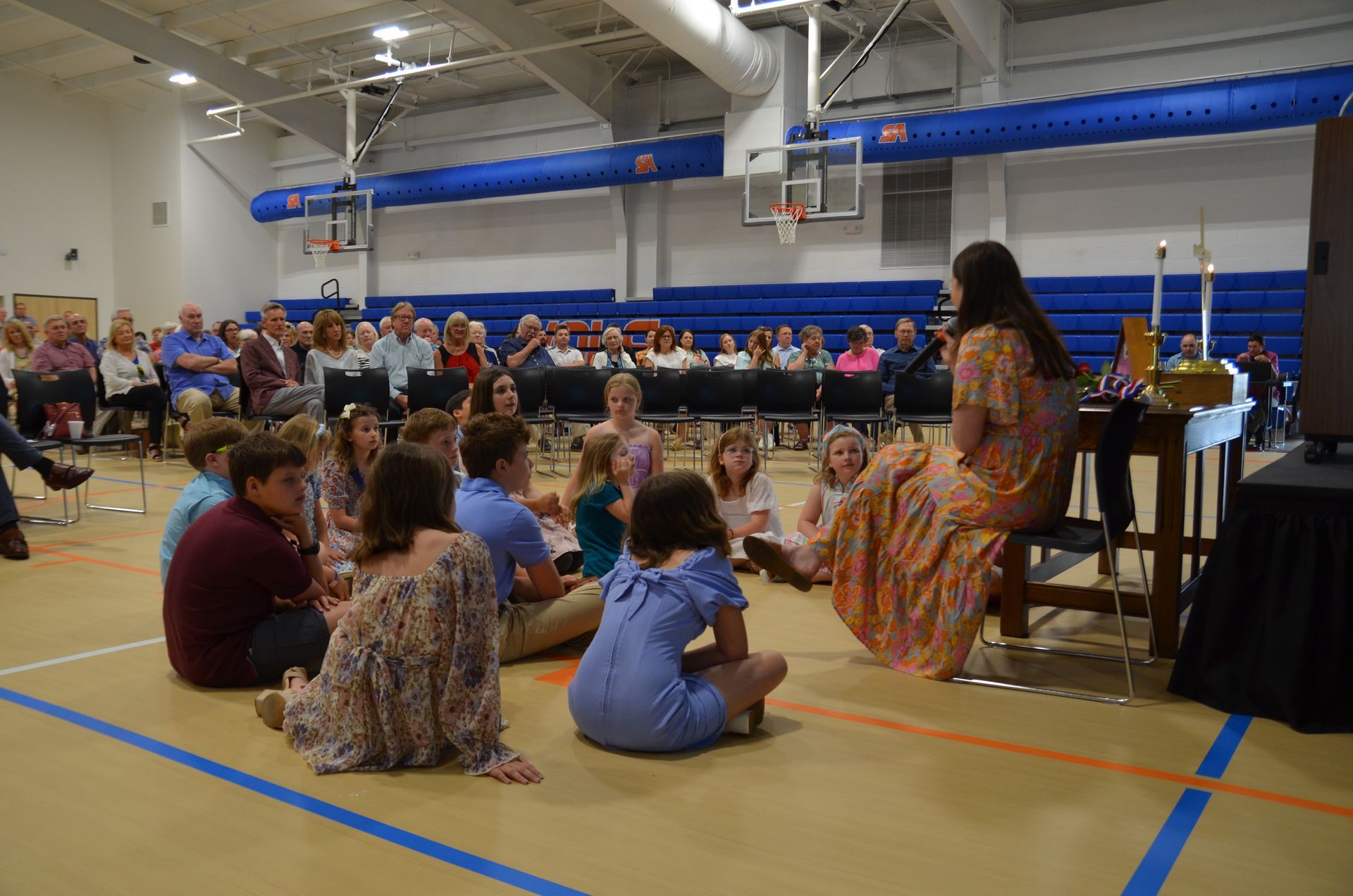 A woman is sitting on a stool in front of a crowd of people in a gym.