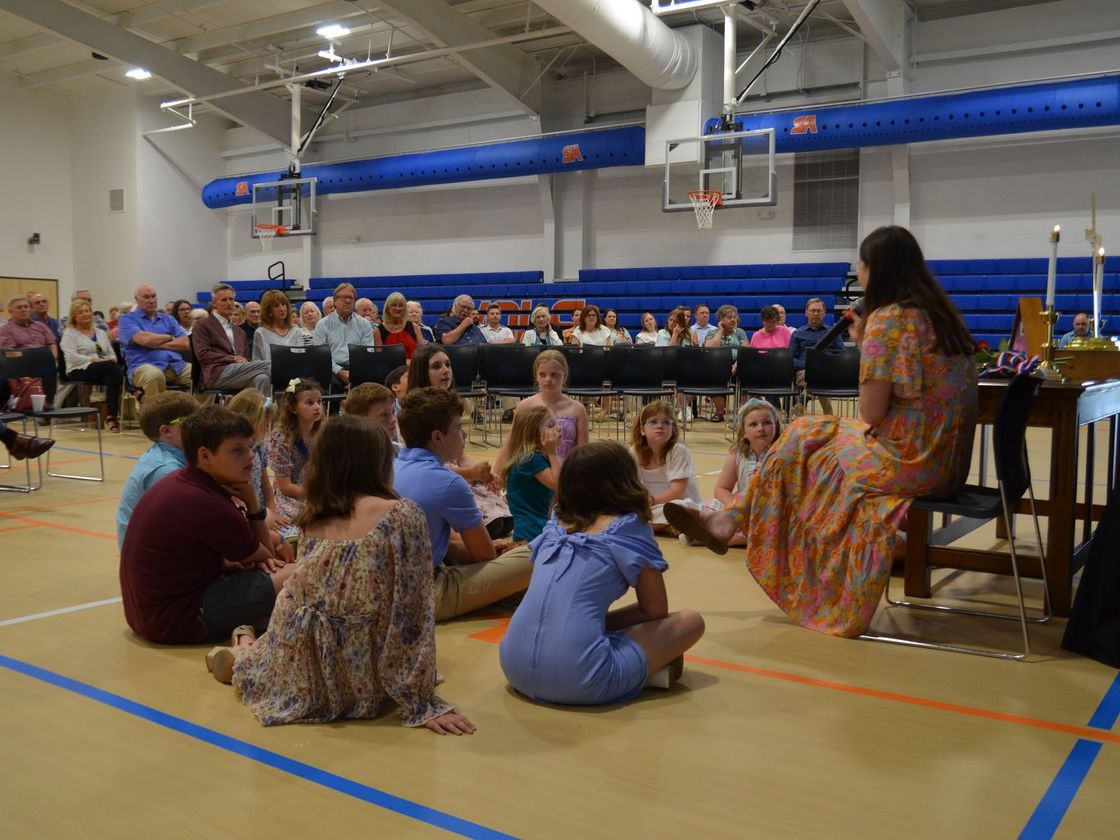 A woman is sitting on a stool in front of a group of children in a gym.