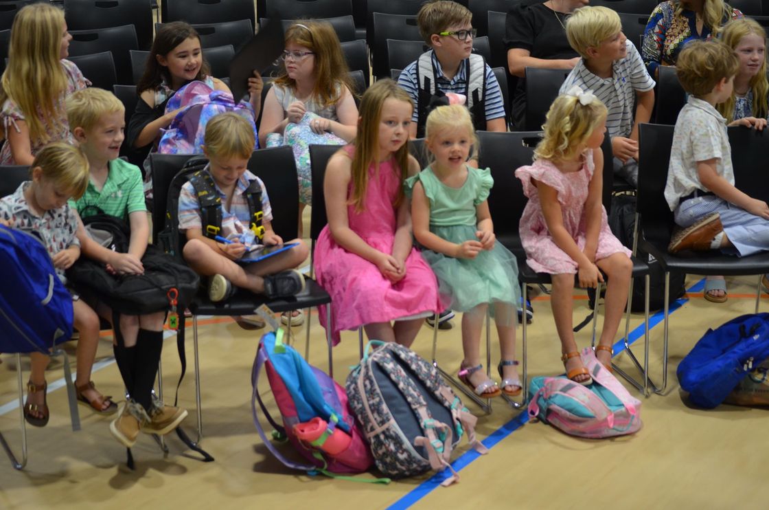 A group of children are sitting in chairs with their backpacks on the floor.