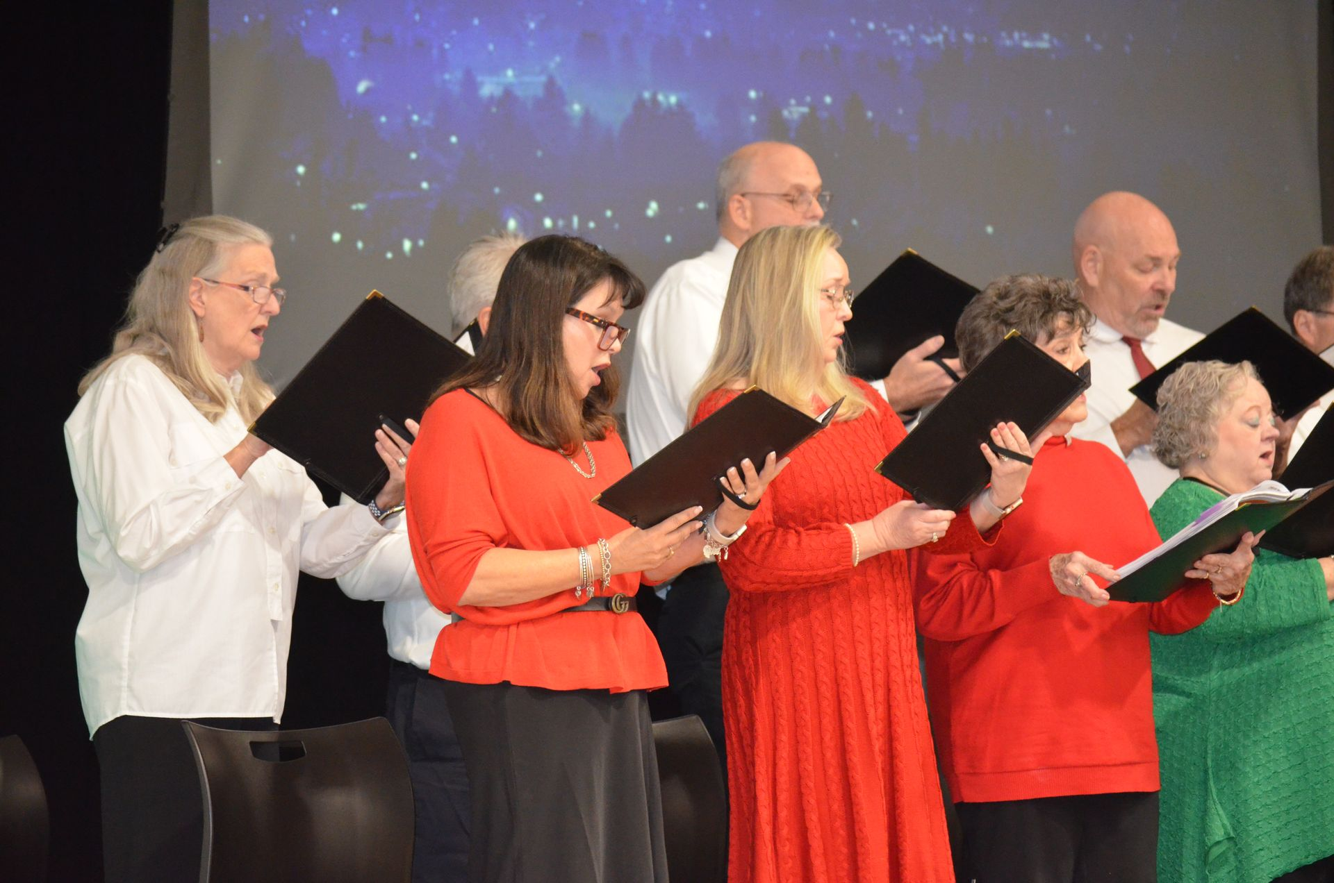 Choir singing with sheet music, stage background with lights, some wearing red tops.