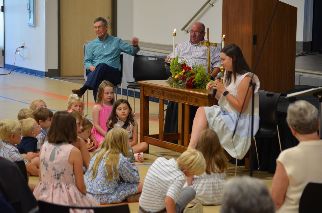 A woman is sitting at a table talking to a group of children.