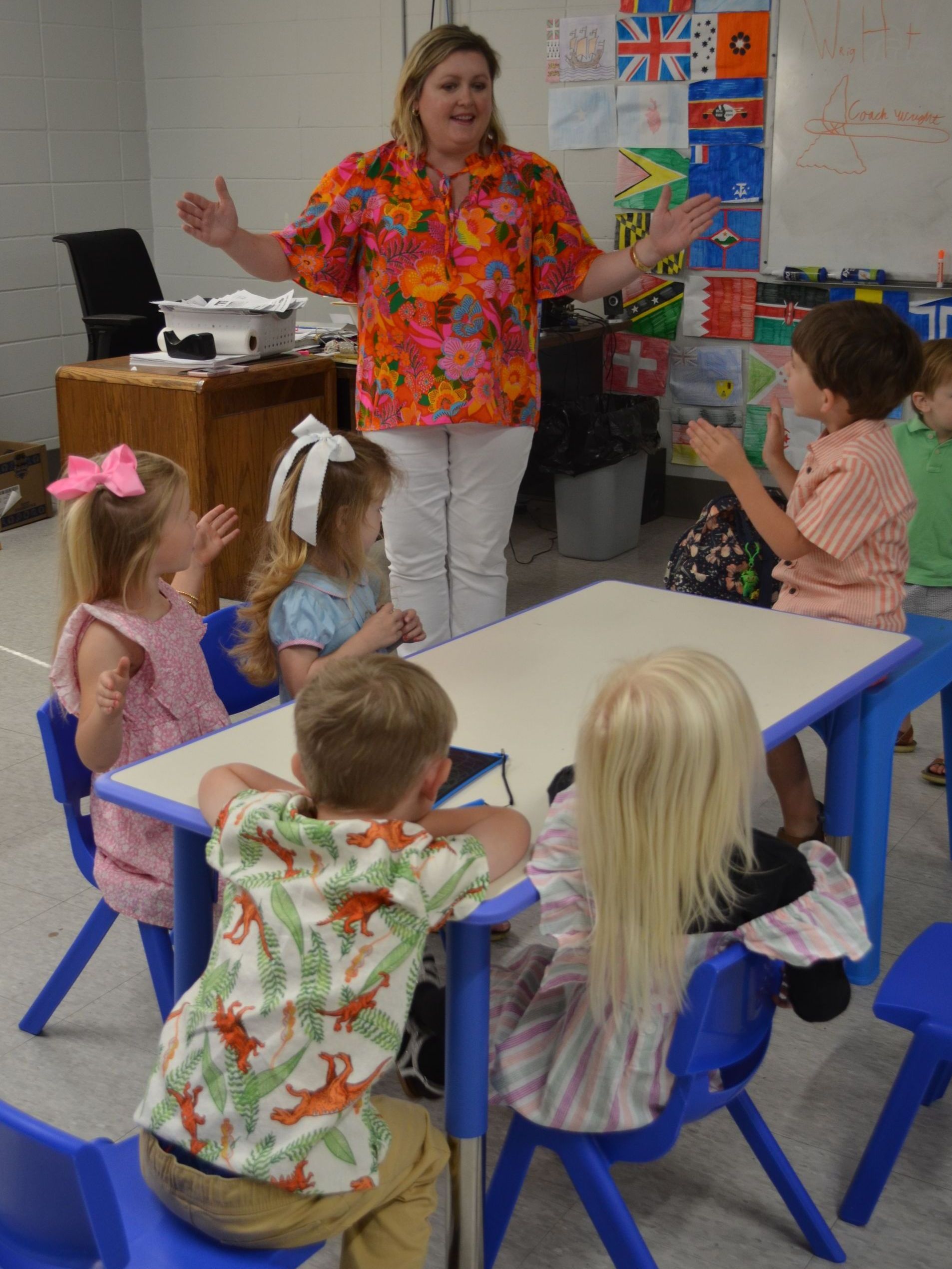 A woman stands in front of a group of children sitting at tables
