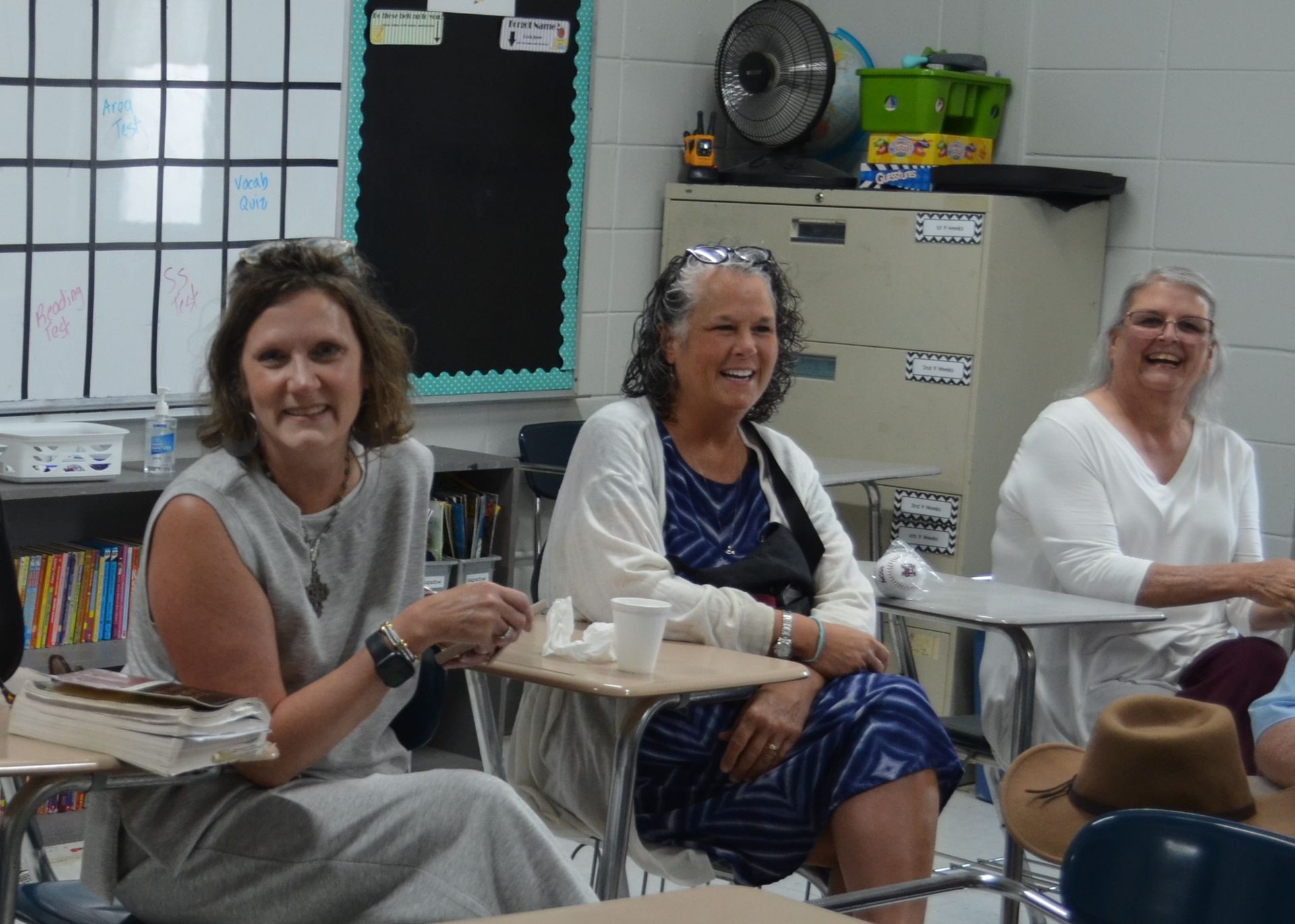 Three women are sitting at desks in a classroom and smiling.