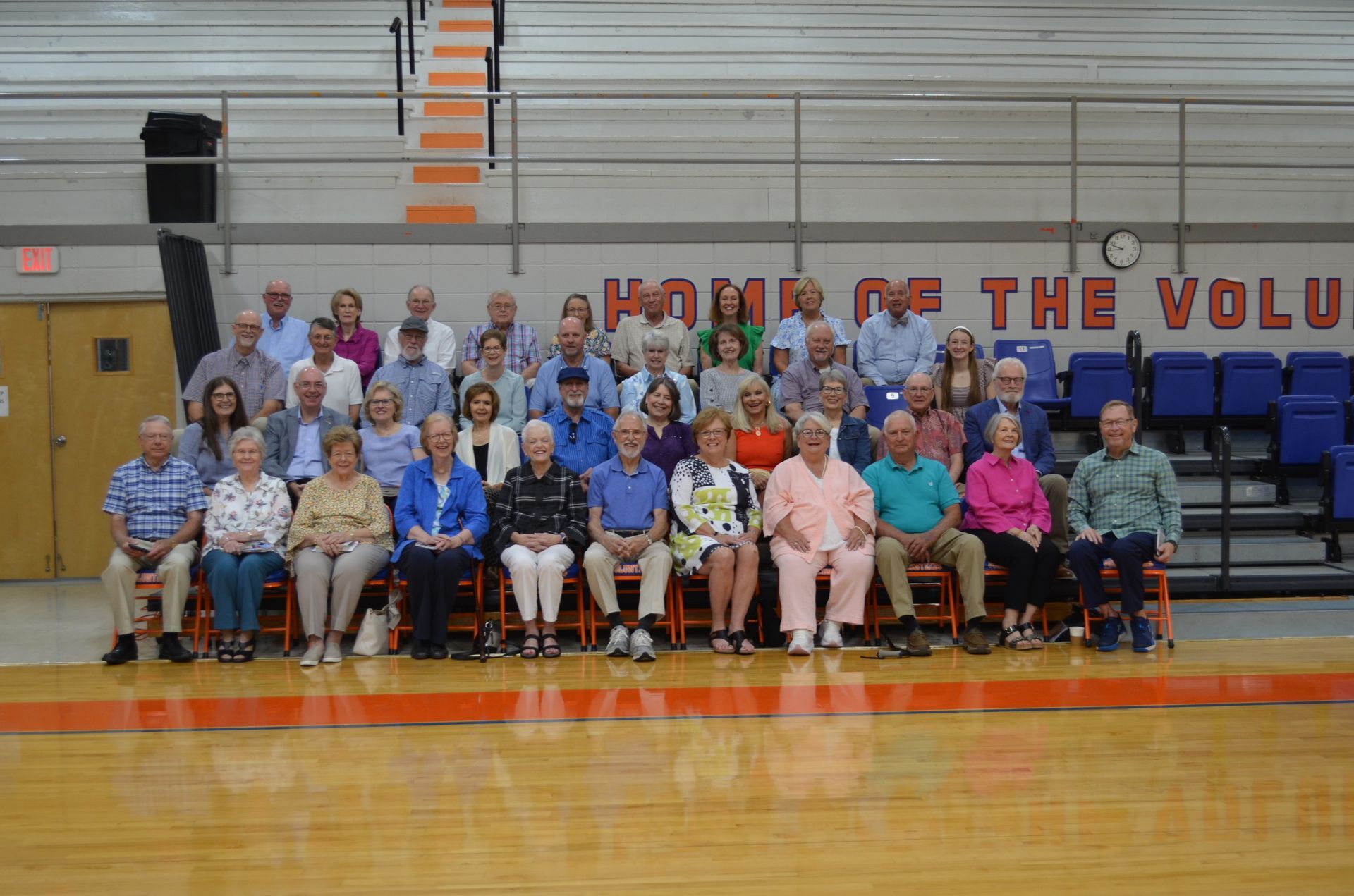 Group photo of people seated and standing in a gymnasium with orange and blue accents, facing the camera.