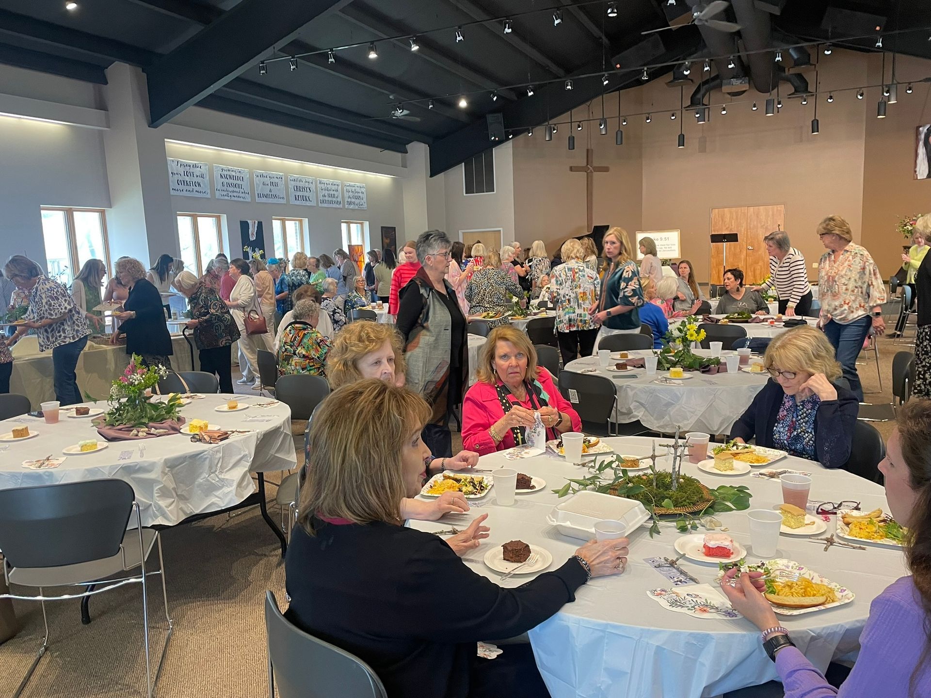 Large room with round tables set for a luncheon, many women gathered, eating and socializing.