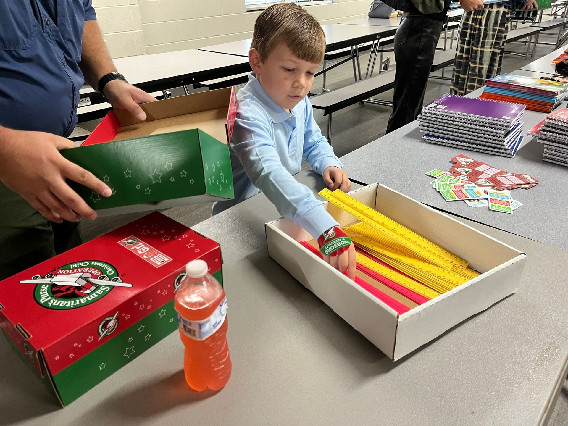Boy placing items in a box, with a man assisting. Boxes are decorated for the holidays; orange drink on table.