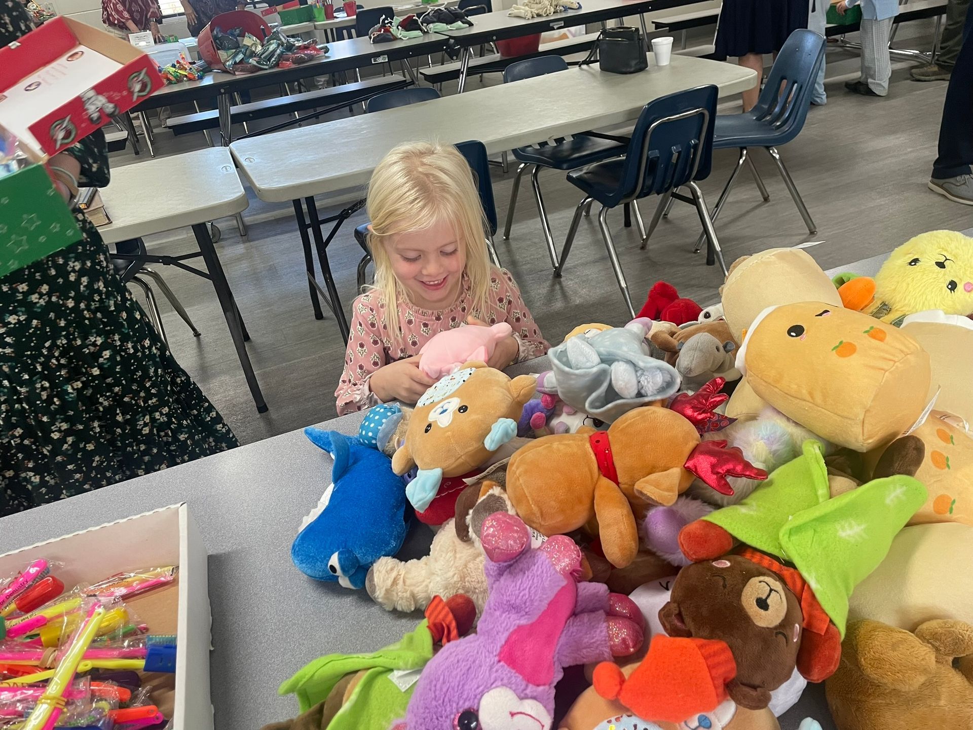 A young child smiles, surrounded by a pile of colorful stuffed animals at a table.