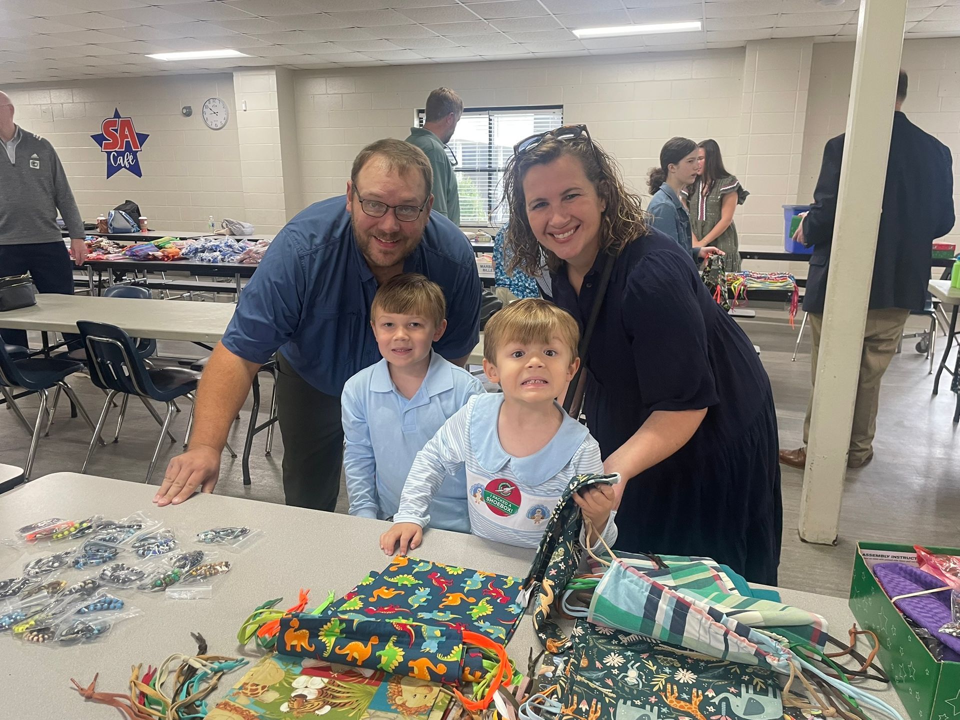 Family at a craft table, smiling. Two young boys, mom, and dad pose with colorful fabric. Indoor setting.