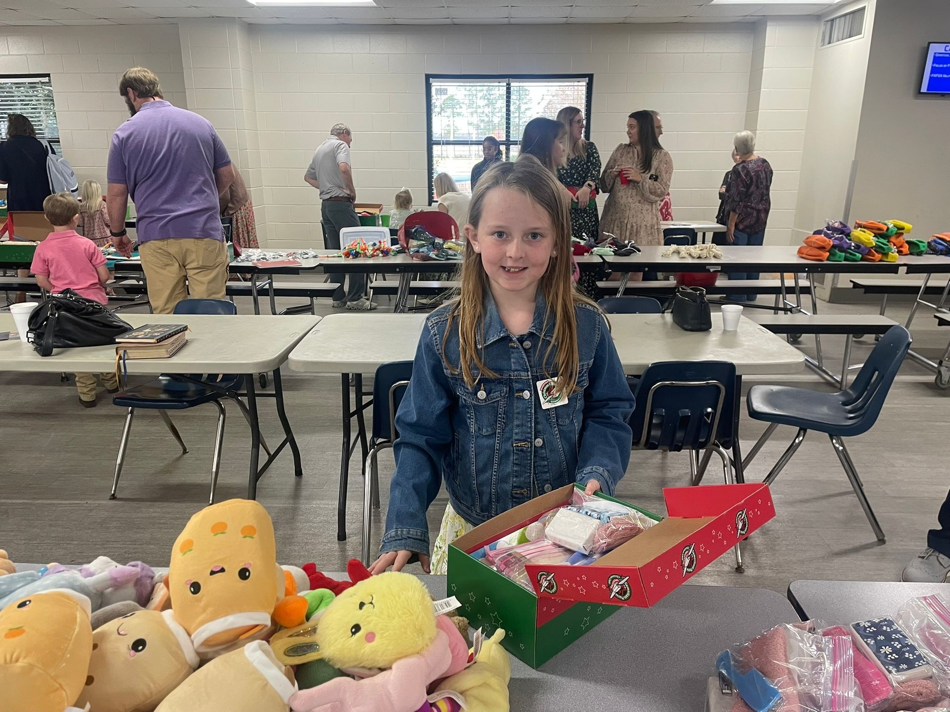 Girl smiles, holding a box of gifts at a donation event. Other people, tables, and toys are visible.