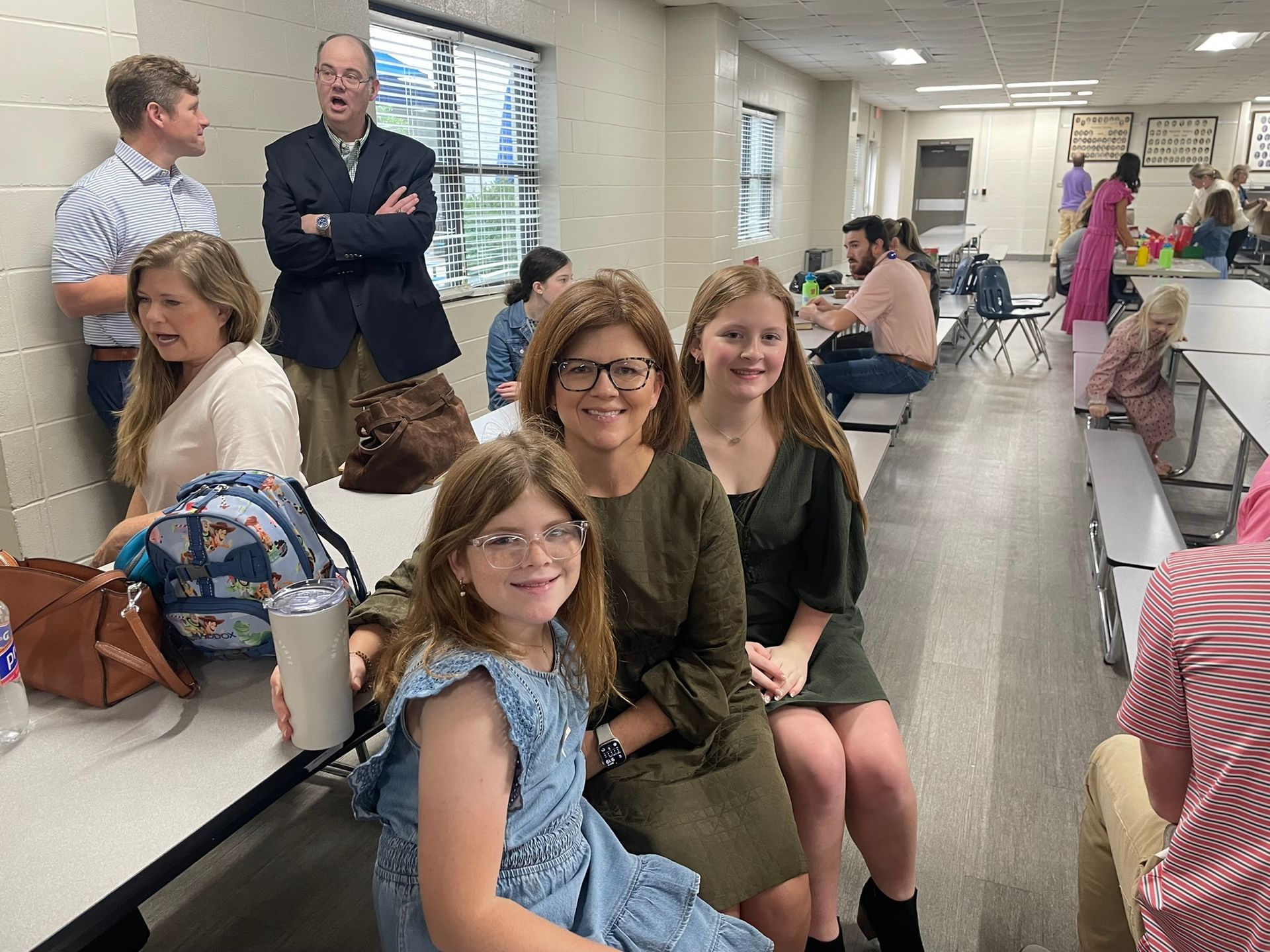 People sitting at tables in a school cafeteria. Woman smiles with two girls, others nearby.