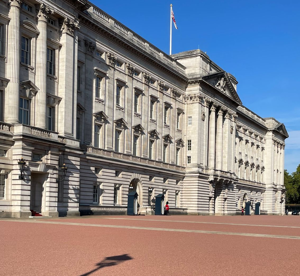 Buckingham Palace facade with Union Jack flag; guards on duty, red brick plaza.