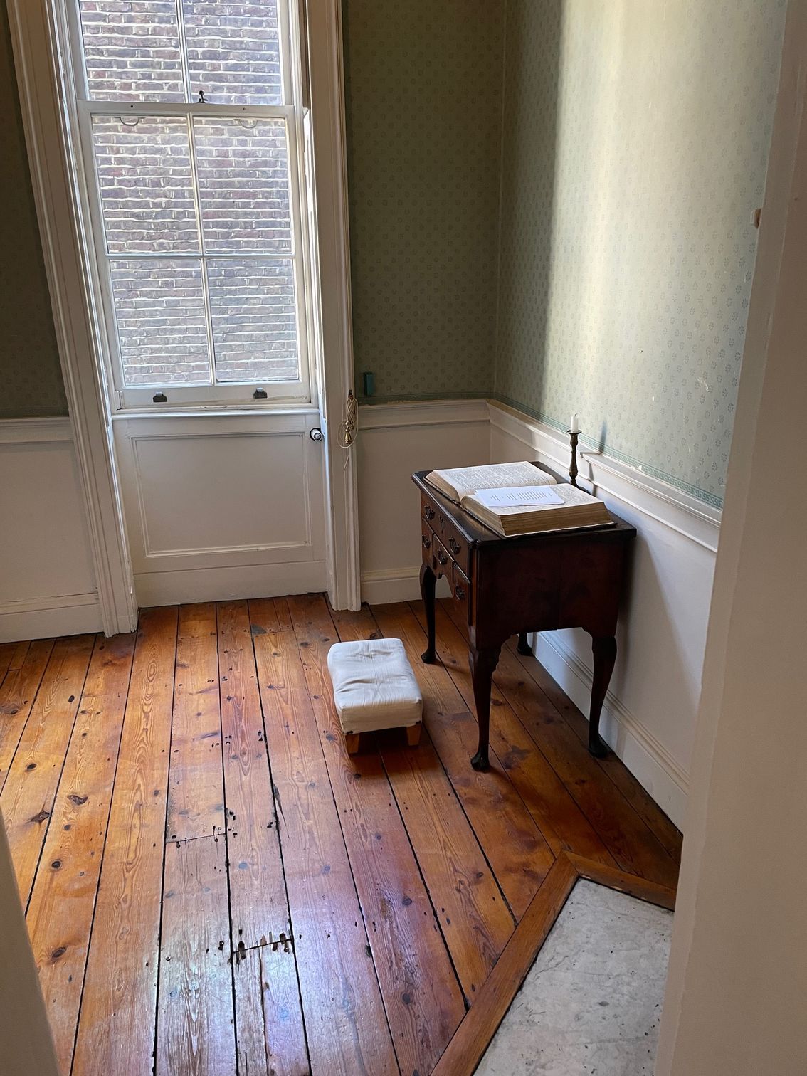 A room with wooden floor, window, and small table. A small stool is on the floor. Walls are painted light green.