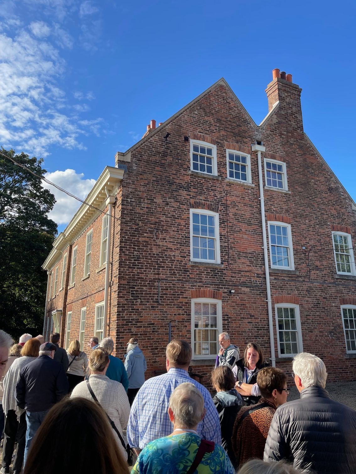Large brick building, several stories, with many windows. A crowd of people stand in front, with blue sky above.
