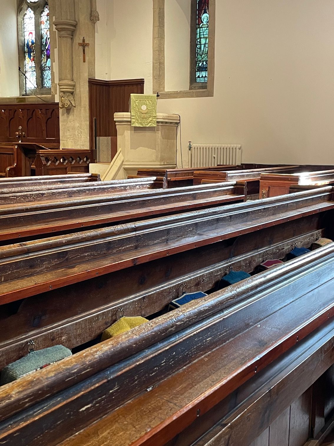 Rows of wooden church pews lead to a stone altar with a green cloth. Stained glass windows and a cross are visible.