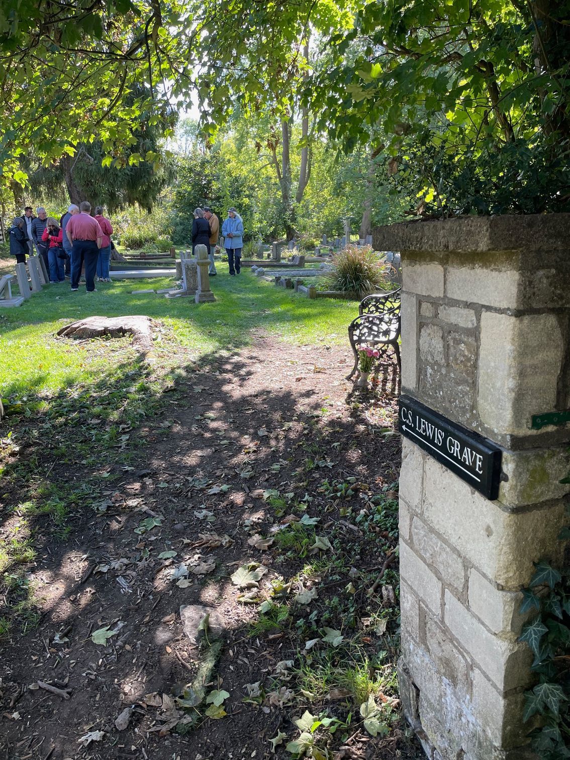 Cemetery scene with path, gravestones, people in the background, and sign on a stone pillar.