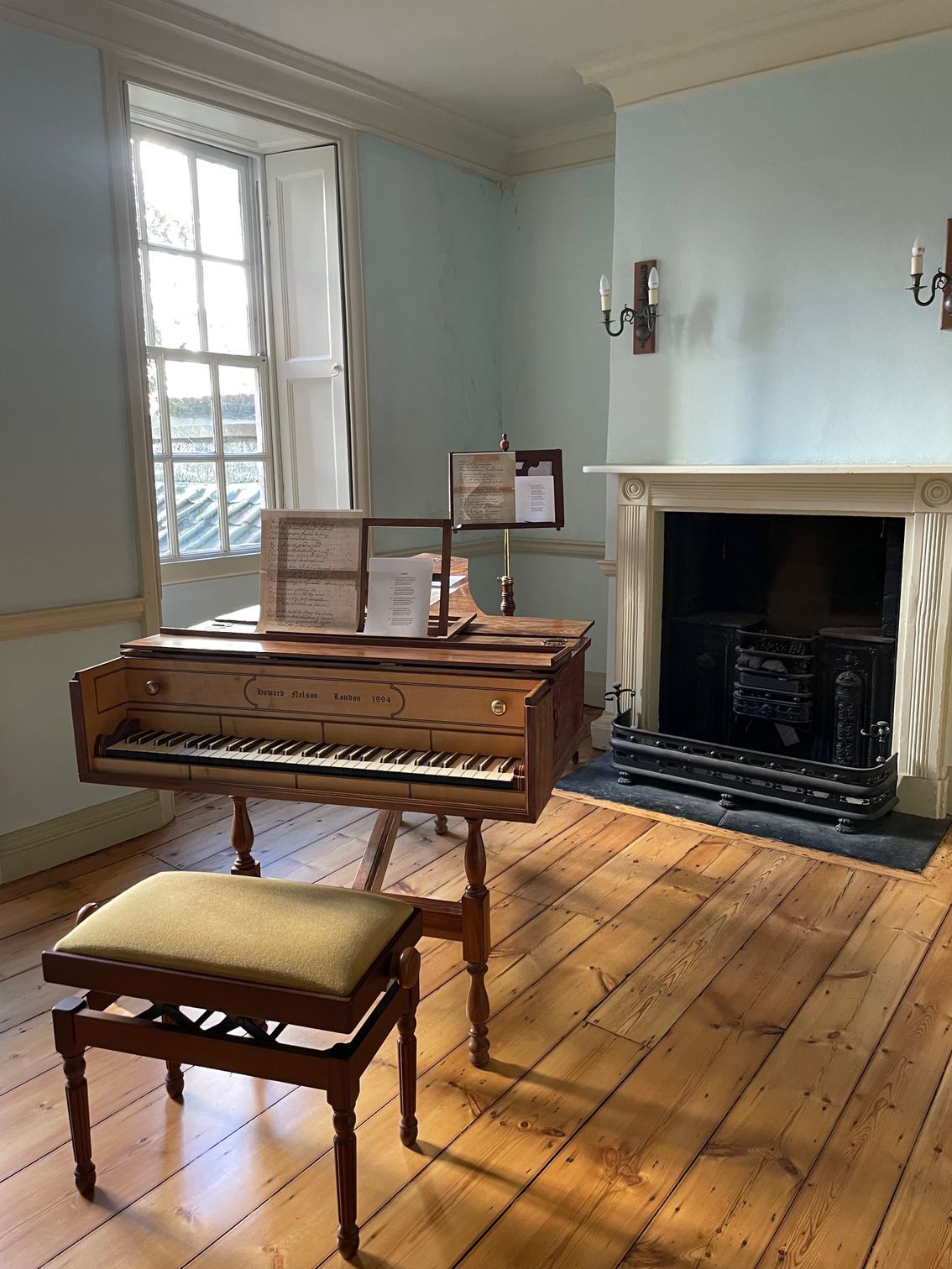 Antique harpsichord and stool in a light blue room with fireplace and window. Wooden floor.
