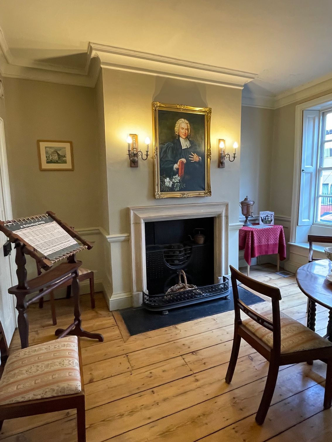 Historical room interior with portrait over fireplace, easel, and period furniture. Wooden floor and neutral walls.