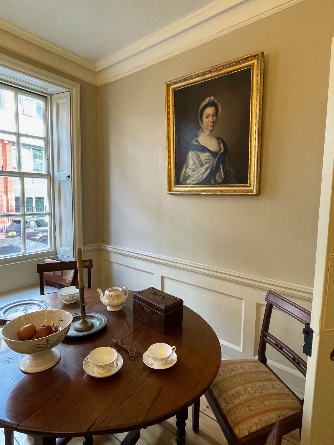 A dining room with a round wooden table, chairs, and portrait. Window on the left, ornate molding.
