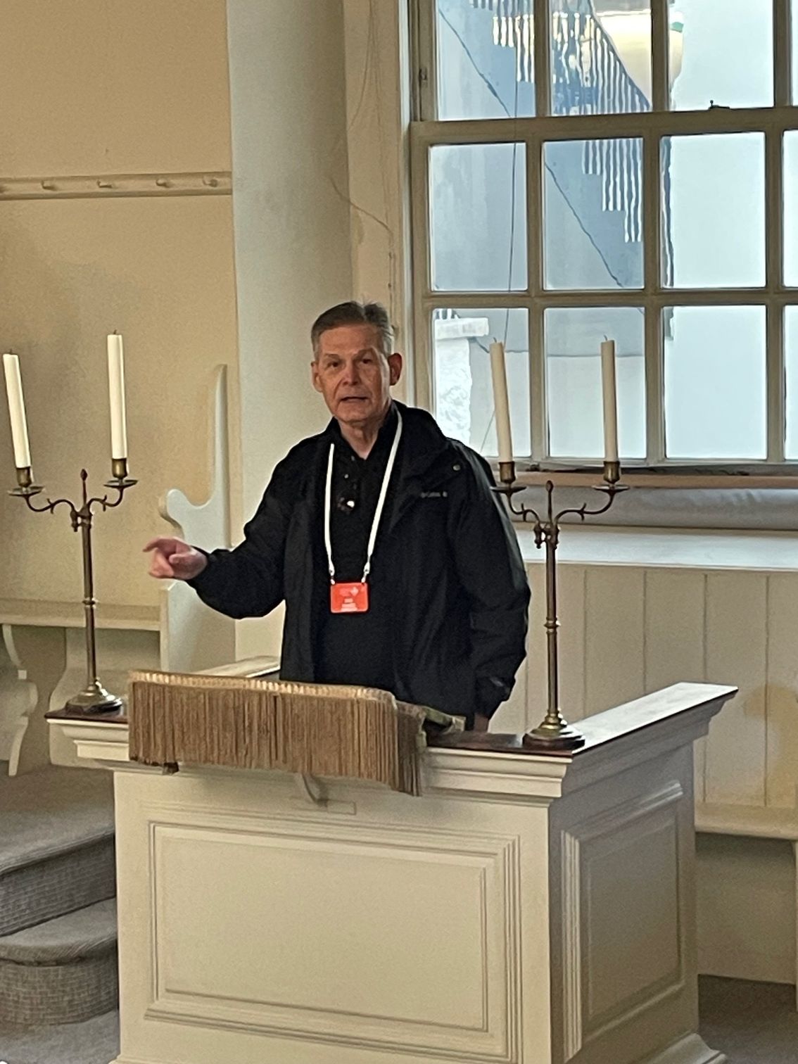 Man speaking from a pulpit in a light-filled room. He gestures, with candles and old books on the pulpit.