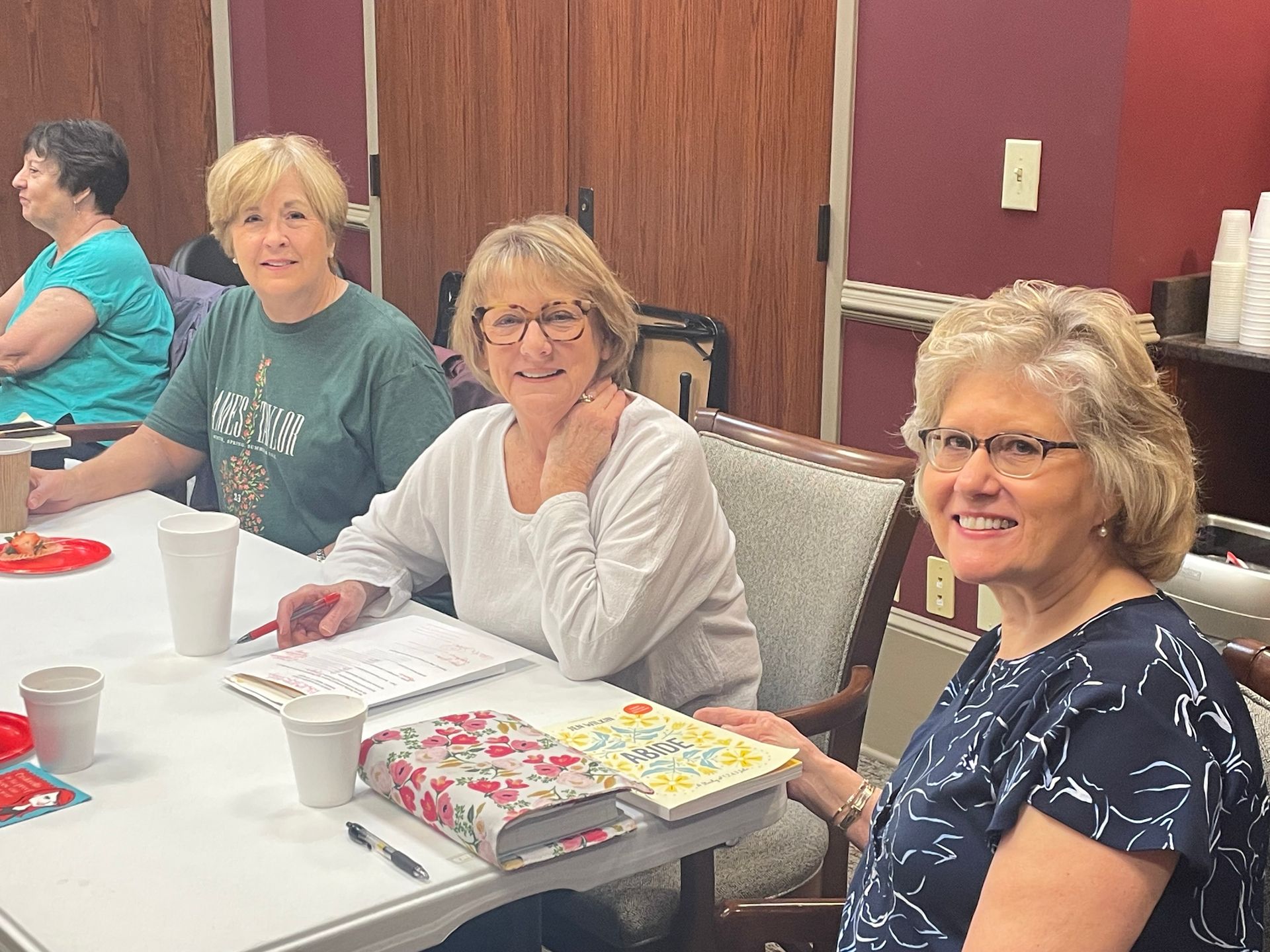 Four women seated at a table, smiling. Notebooks and cups are visible. Wooden paneling in the background.