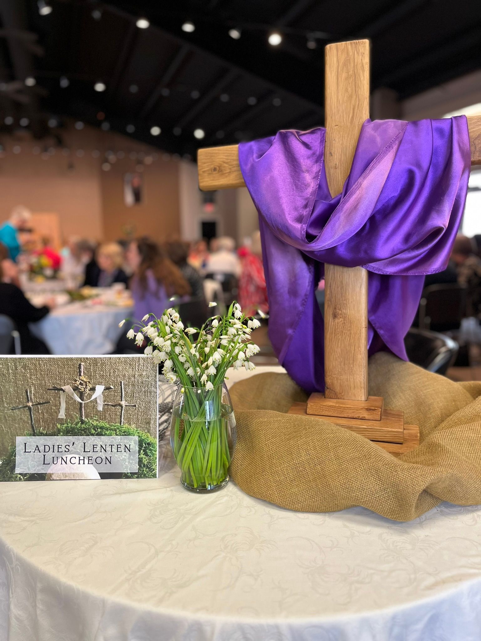 Wooden cross with purple cloth draped over it, on a table. Flowers and a sign are nearby.