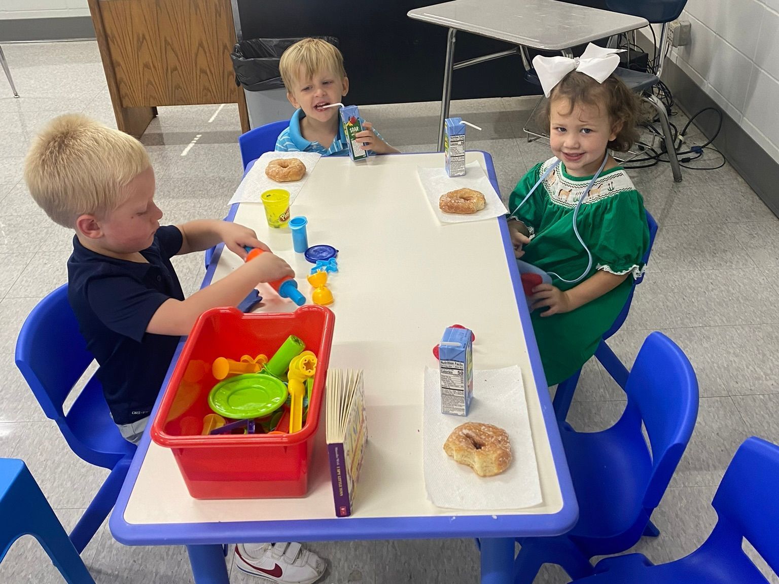Three children are sitting at a table playing with toys and donuts.
