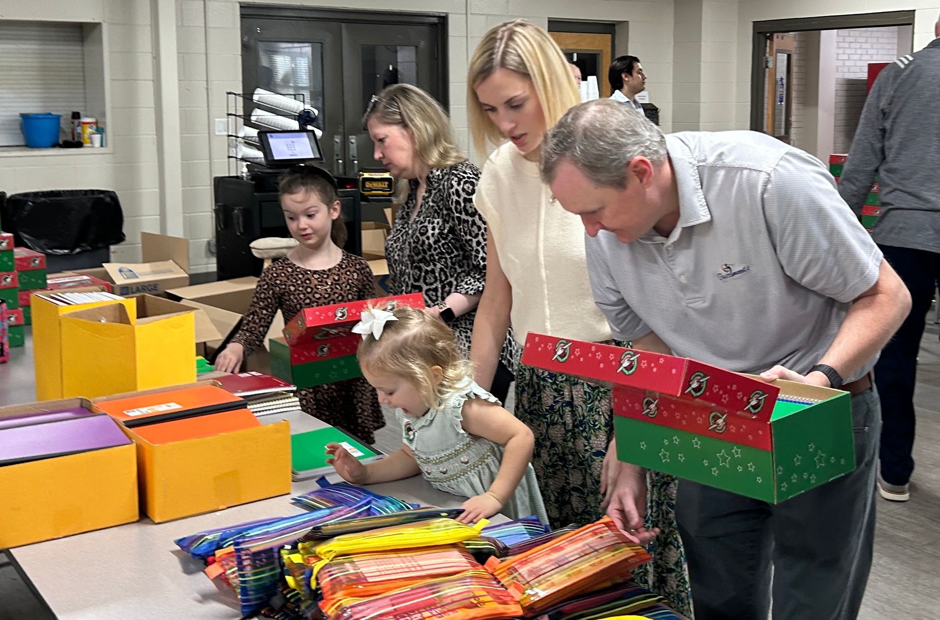 People packing shoeboxes at a table. Two children and three adults inspect gifts. Indoors, festive boxes and supplies are visible.