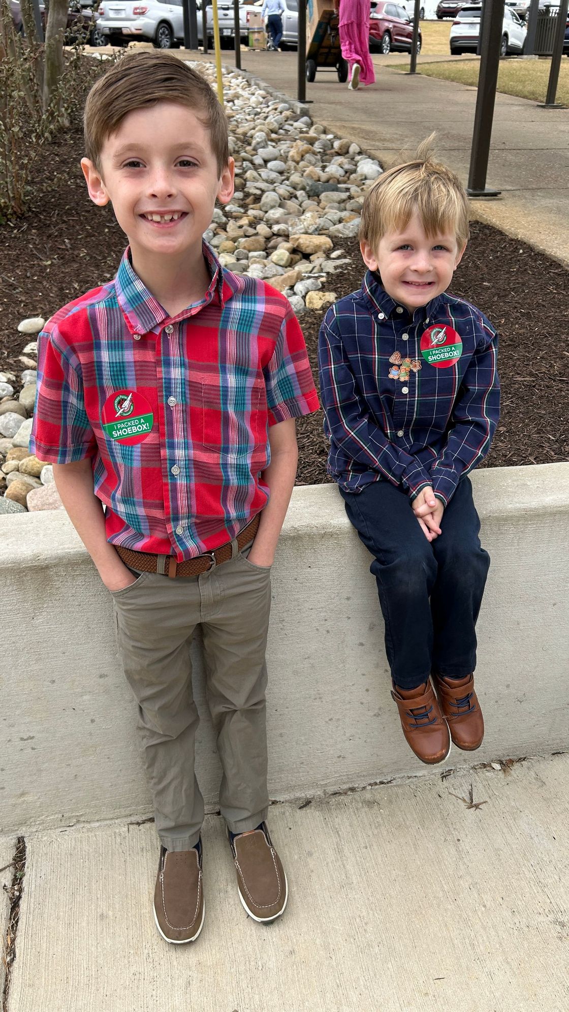 Two young boys pose outdoors: one standing, one seated. The standing boy wears a red plaid shirt, the seated boy a blue sweater.