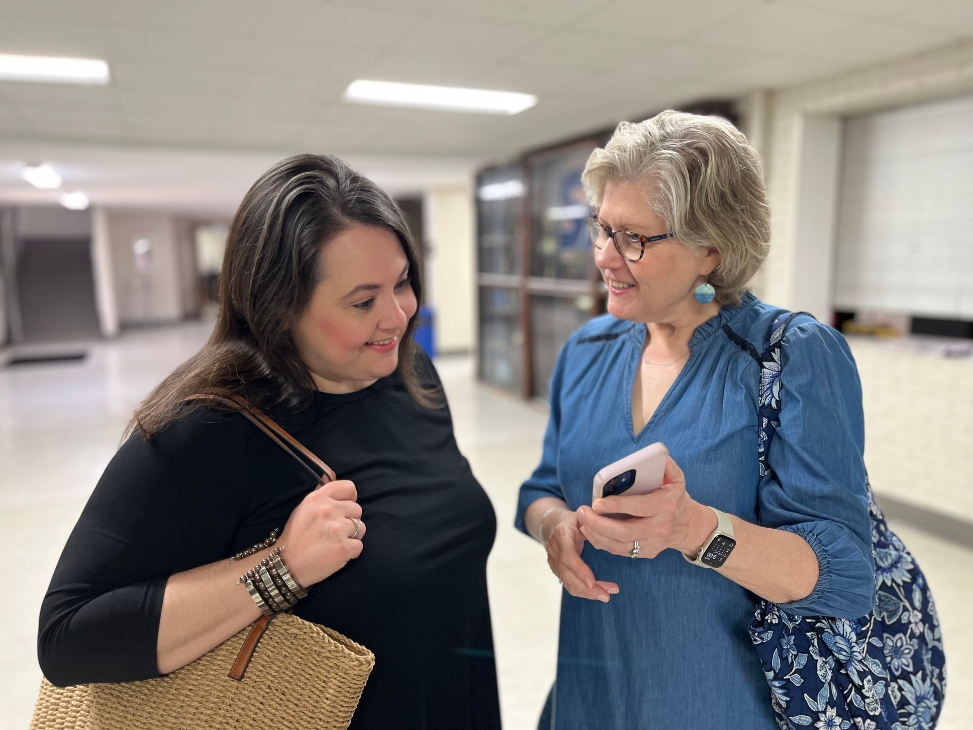 Two women conversing in a hallway. One holds a phone, the other a straw tote bag. Soft lighting.