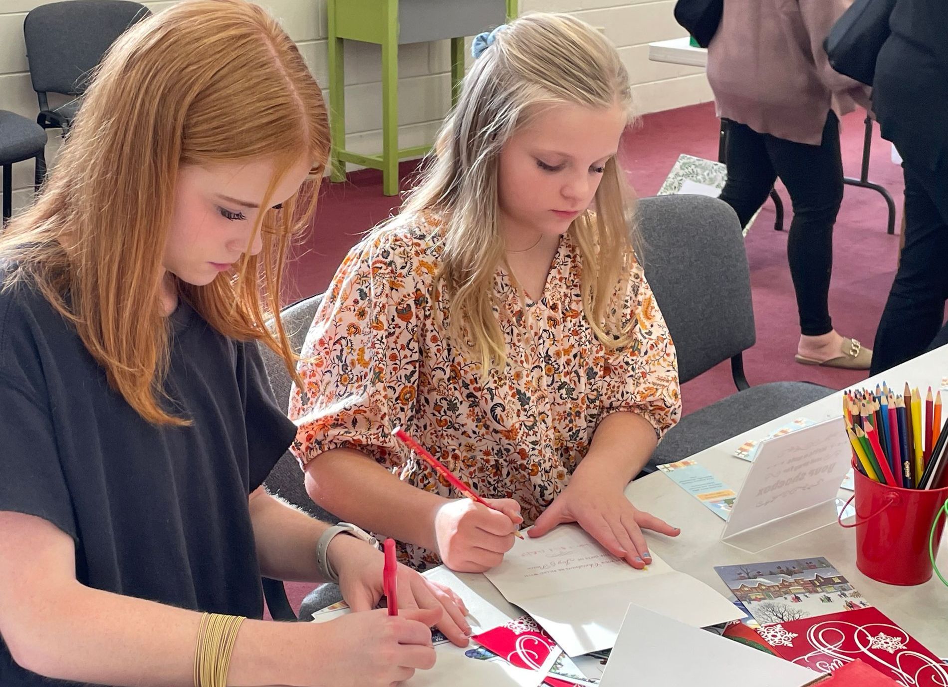 Two young girls are sitting at a table writing on a piece of paper.