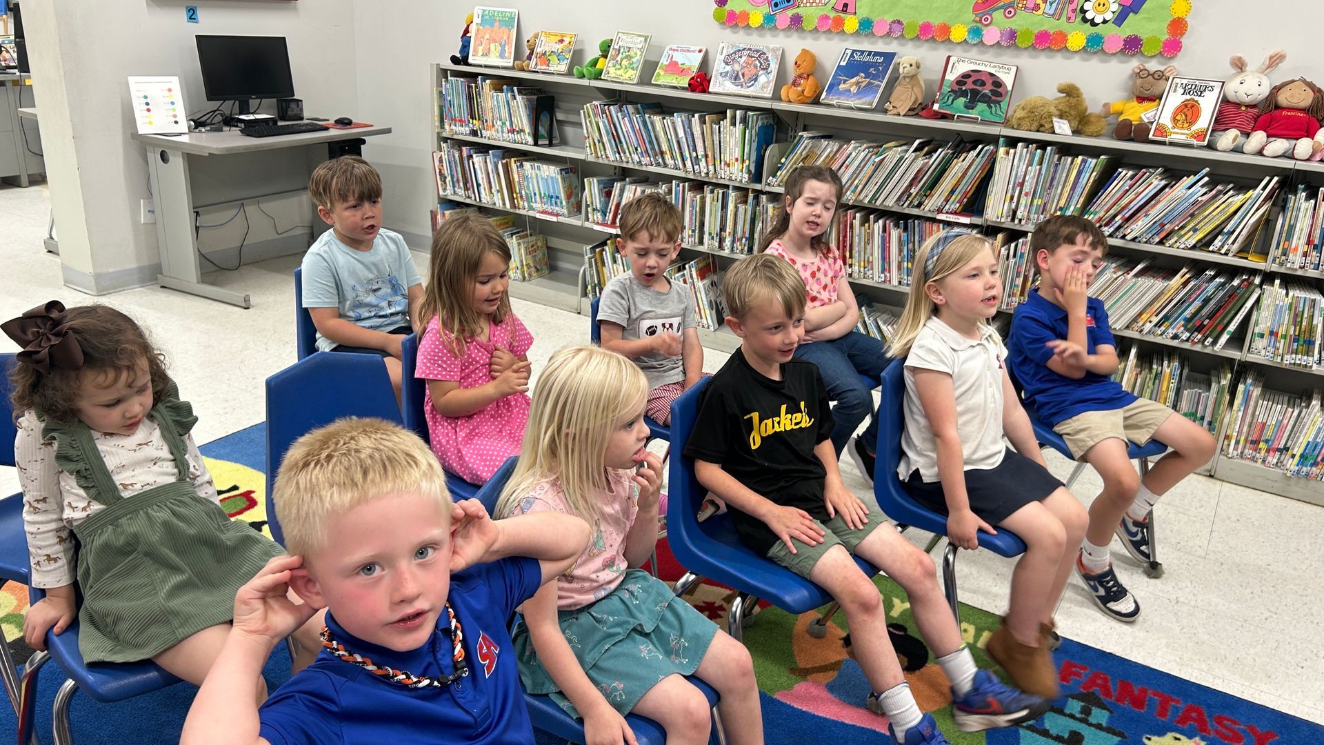 Children sitting in blue chairs, listening in a library. Bookshelves are in the background.