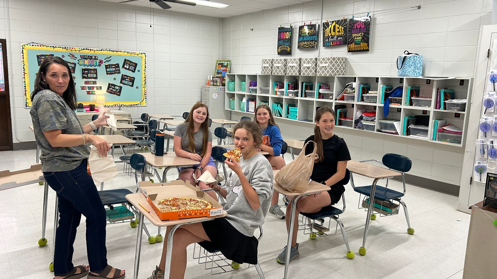 A teacher and students in a classroom with pizza and lunch bags.