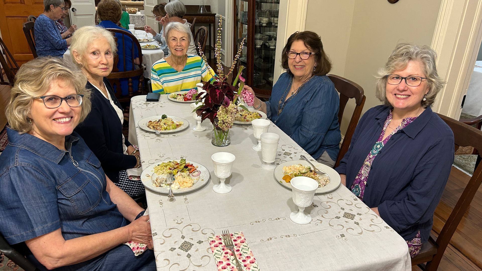 Six women sit at a decorated table, smiling. There are plates of food and flowers on the table.