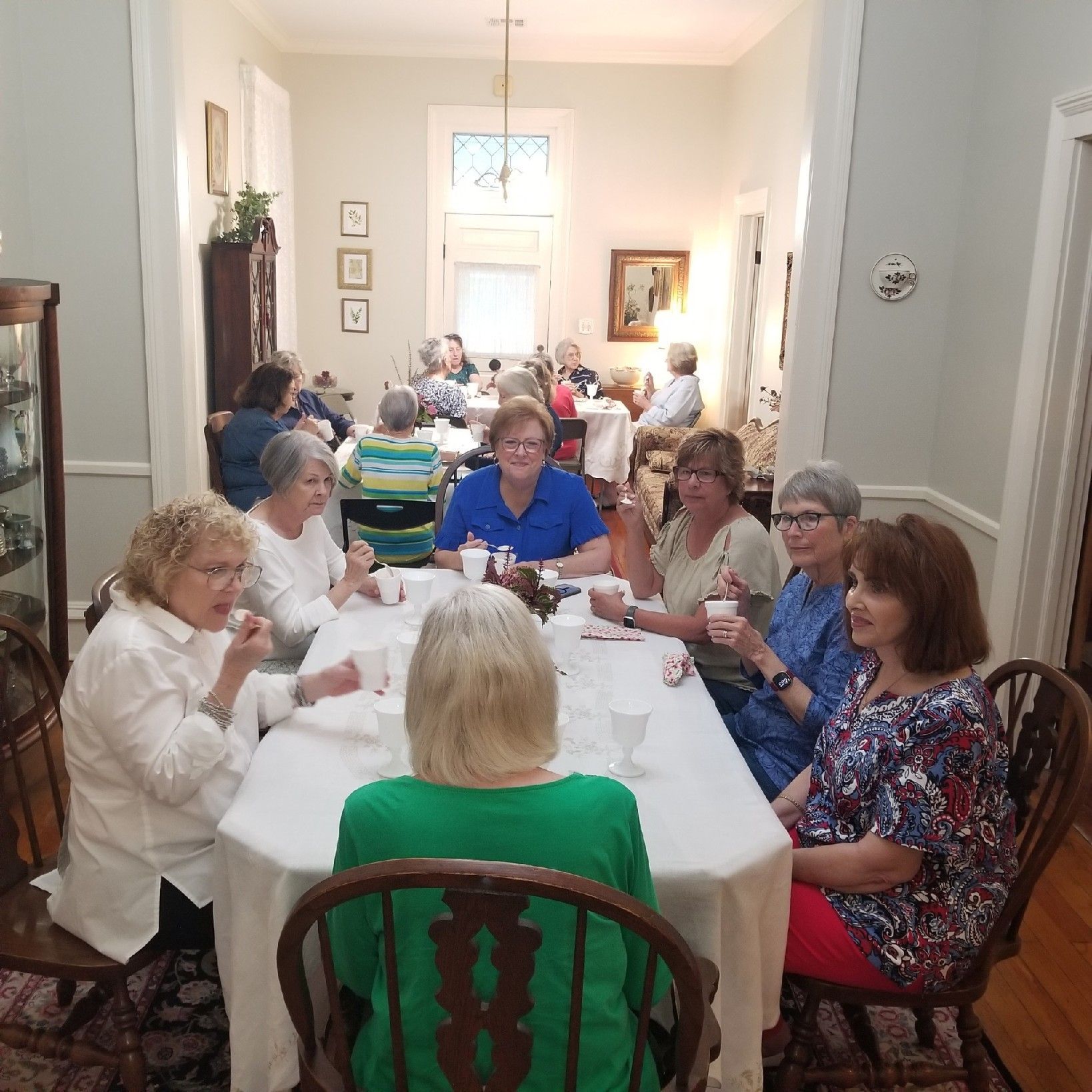 Group of women at a long table, indoors, socializing while having tea or a meal.