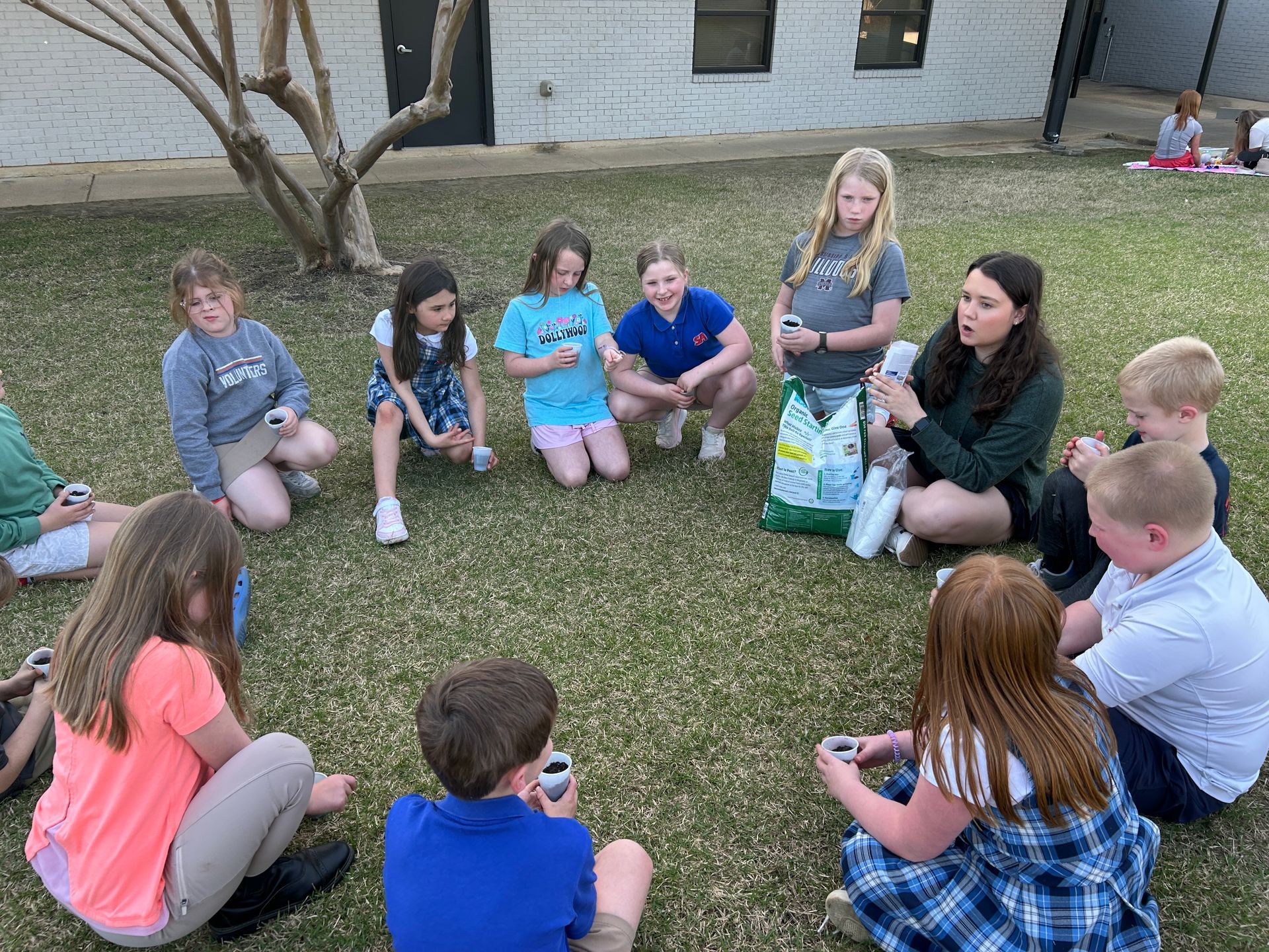 Children in a circle listening to a woman showing them garden supplies on grassy ground.