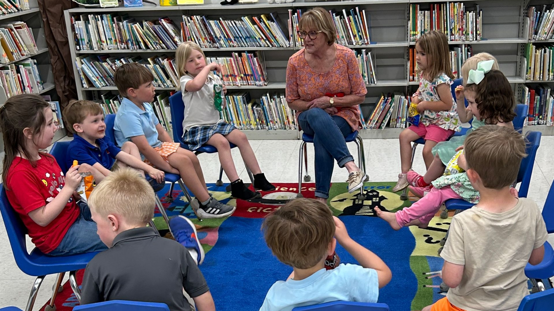 Children seated in a circle listening to a woman reading in a library.