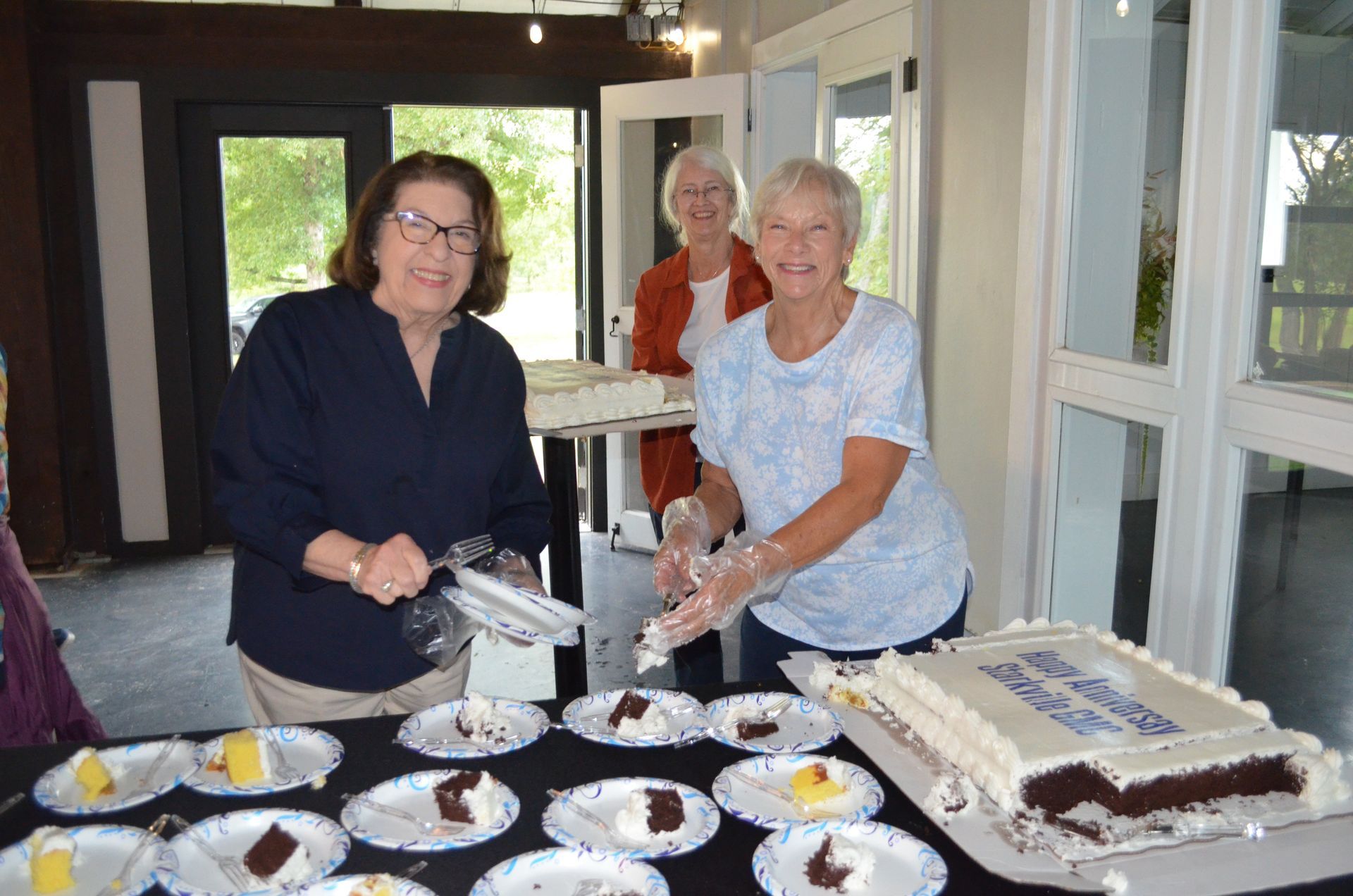 Three women cut and serve cake at an event. White frosted cake sits on a table, ready to be served.